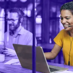 Frauen mit Headset lächelt, während sie auf einem Laptop arbeitet. Im Hintergrund ein Mann, der ebenfalls am Laptop sitzt. Beide nutzen eine UC-Plattform.