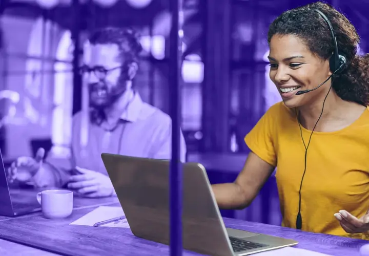 Frauen mit Headset lächelt, während sie auf einem Laptop arbeitet. Im Hintergrund ein Mann, der ebenfalls am Laptop sitzt. Beide nutzen eine UC-Plattform.