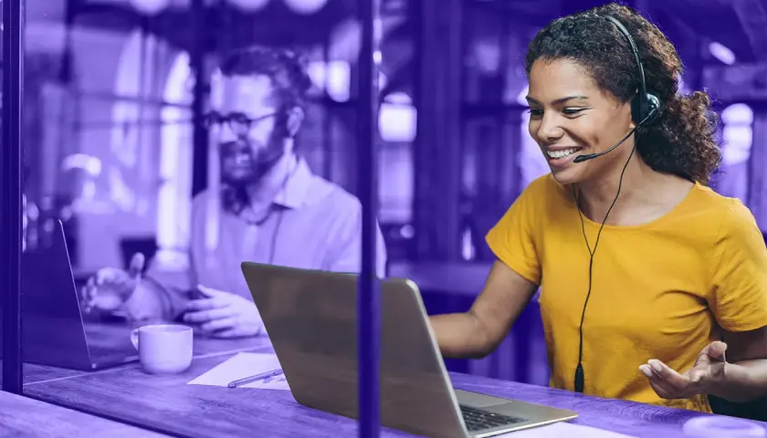Frauen mit Headset lächelt, während sie auf einem Laptop arbeitet. Im Hintergrund ein Mann, der ebenfalls am Laptop sitzt. Beide nutzen eine UC-Plattform.